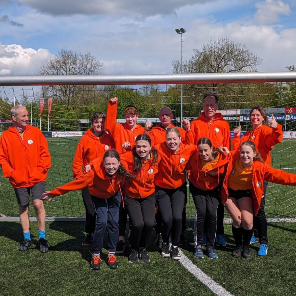 Elf scheidsrechters in oranje hoodies, jong en oud door elkaar, juichen voor een voetbaldoel op het kunstgrasveld.